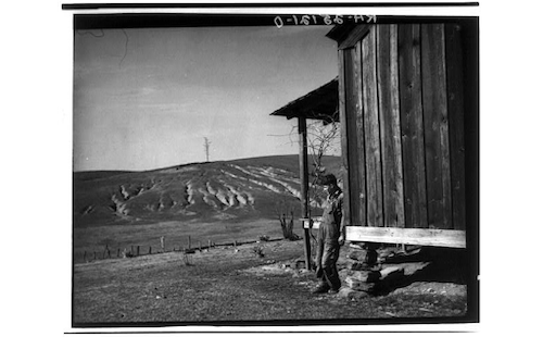 On the right side of the photo is a wooden shack on a tenant farm during the Great Depression. A farmer in overalls and a hat leans against it, looking unhappy.  The left side of the photo is a view of a hill with a farm field on it and the sky above it. 
Photographer: Arthur Rothstein, 1915-1985. Source Collection: Farm Security Administration - Office of War Information Photograph Collection (Library of Congress). Library of Congress Digital ID cph 3a01357 //hdl.loc.gov/loc.pnp/cph.3a01357. LCCN Permalink https://lccn.loc.gov/2017775644. Credit Line: Library of Congress, Prints & Photographs Division, Farm Security Administration/Office of War Information Black-and-White Negatives. Public domain via Library of Congress